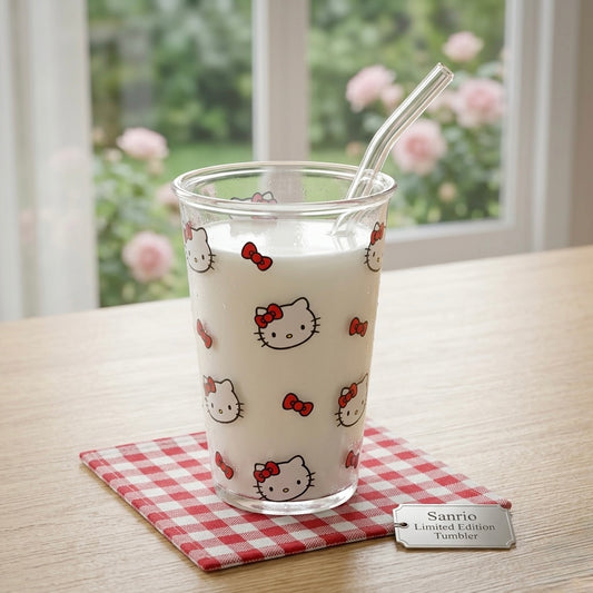 A clear glass tumbler with a Hello Kitty print, filled with milk and a bent glass straw, sits on a red-and-white checkered coaster on a wooden table. A small tag reads 'Sanrio Limited Edition Tumbler.