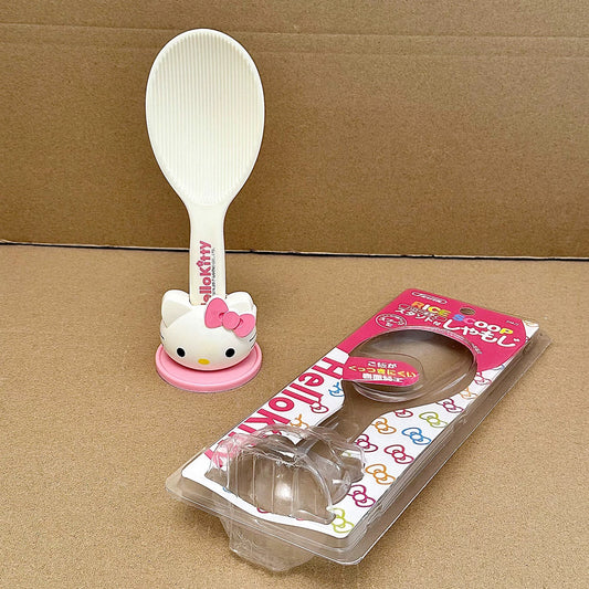 A Hello Kitty plastic spoon with a white handle, pink base, and Hello Kitty character head, shown next to its retail packaging on a brown cardboard background.
