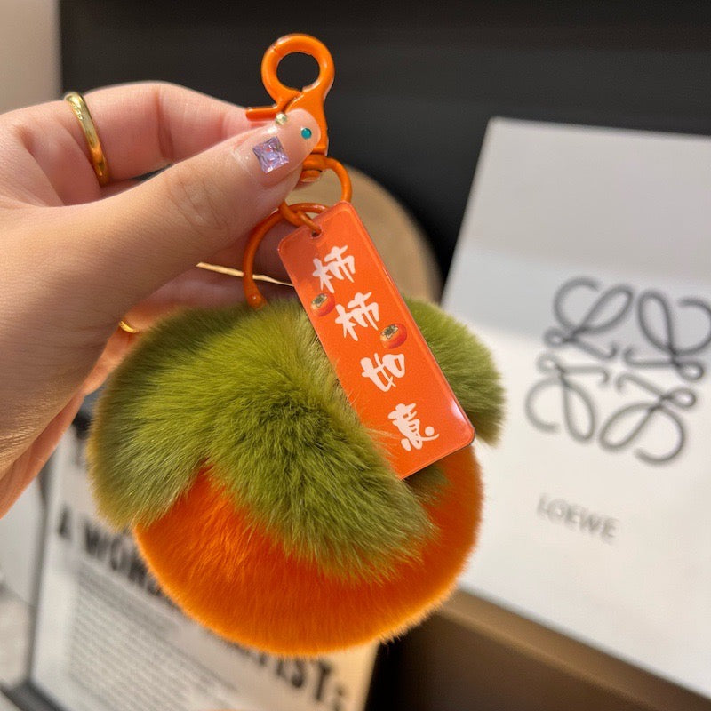 A pair of fluffy persimmon keychains with orange plush bodies and green fuzzy leaves, featuring a lucky charm with the phrase "Good Things Happen" and an orange clip.