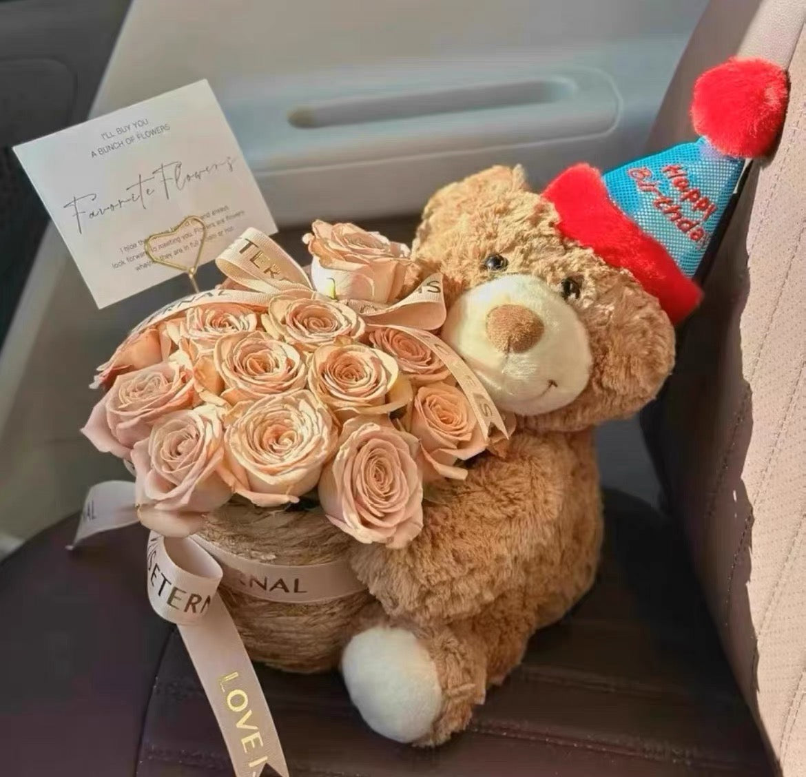 A teddy bear wearing a festive hat sitting next to a bouquet of peach-toned roses in a woven basket, with a 'Happy Birthday' card attached to the side.