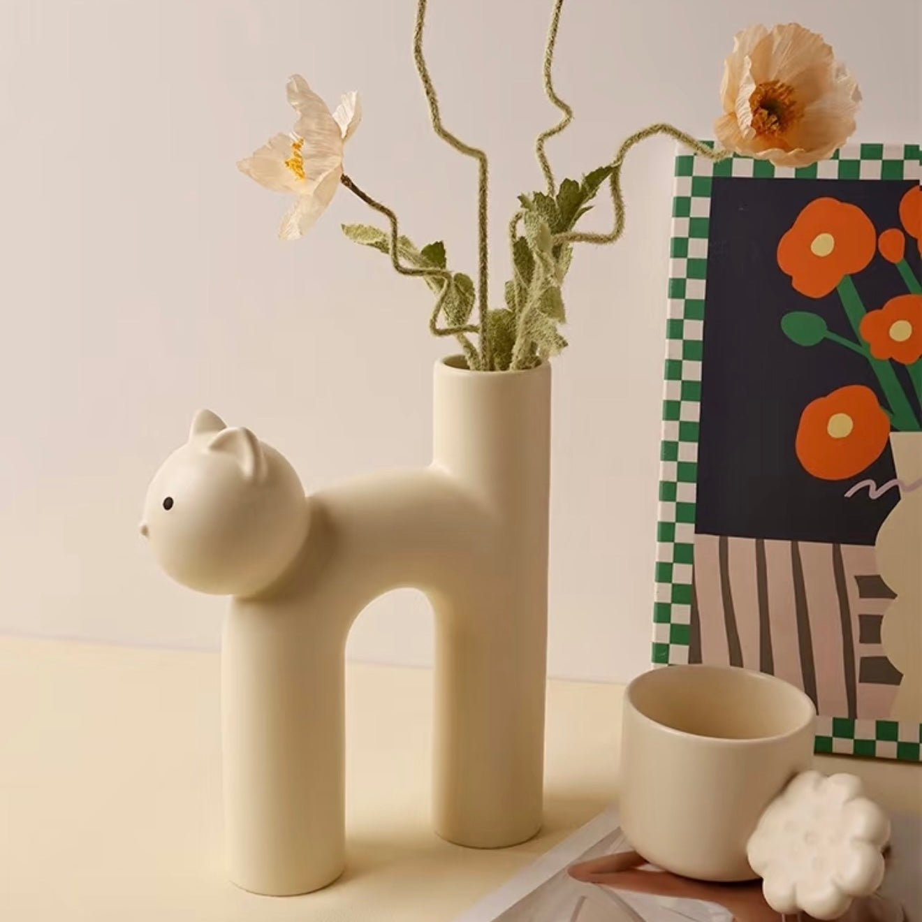 White ceramic vase shaped like a cat with flowers, next to a small white cup and a decorative painting on a light background.