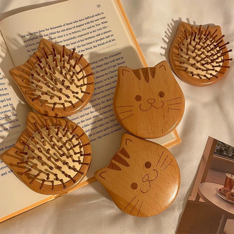 Wooden cat-shaped hair brushes with engraved cat faces, placed on a book and soft fabric background.