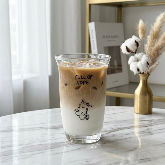 A clear glass cup with black printed text reading 'FULL OF HOPE' and a small cat illustration, filled with an iced coffee drink, set on a marble table in a bright indoor setting.