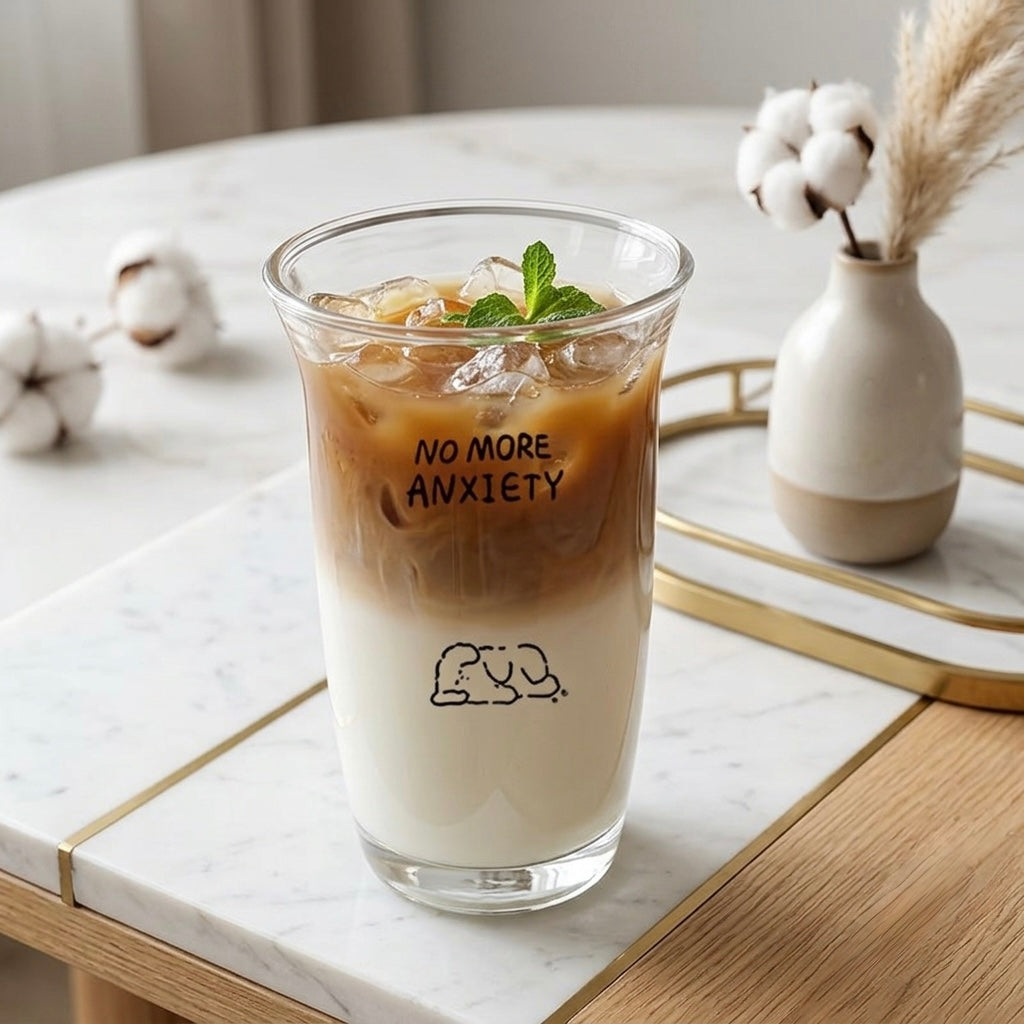 A clear glass cup with black printed text reading 'NO MORE ANXIETY' and a small line drawing, filled with an iced coffee drink and topped with mint, set on a marble table with cotton stems and a small vase in the background.