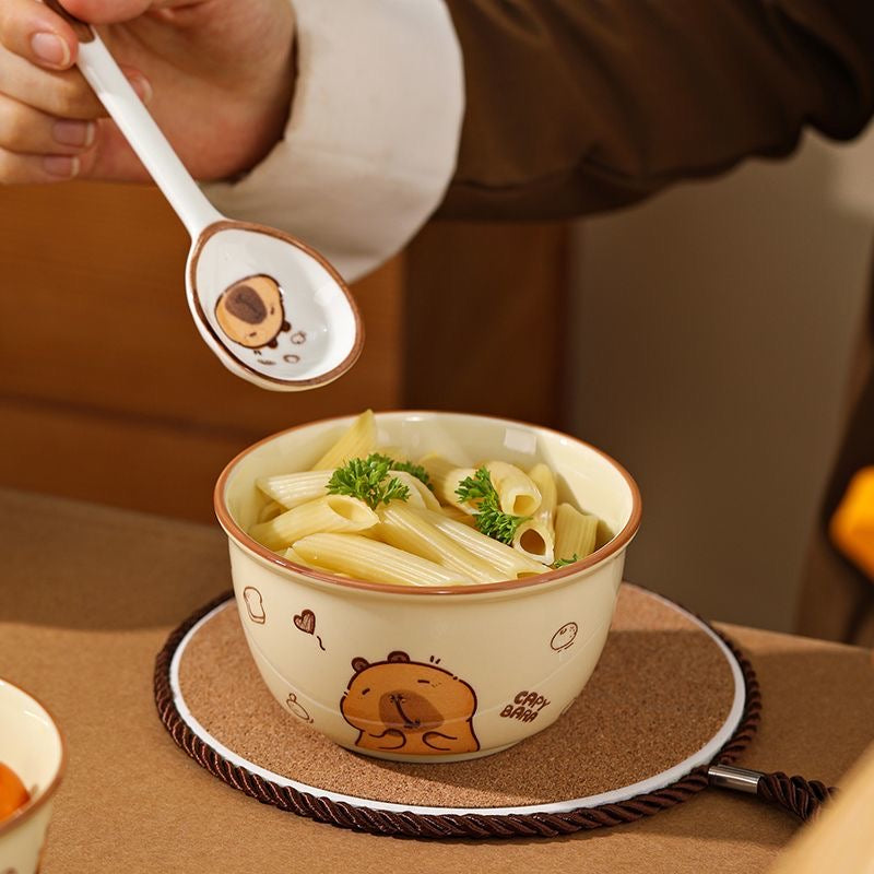 Bowl of pasta with a cartoon bear design on a coaster, accompanied by a spoon with a similar bear design.