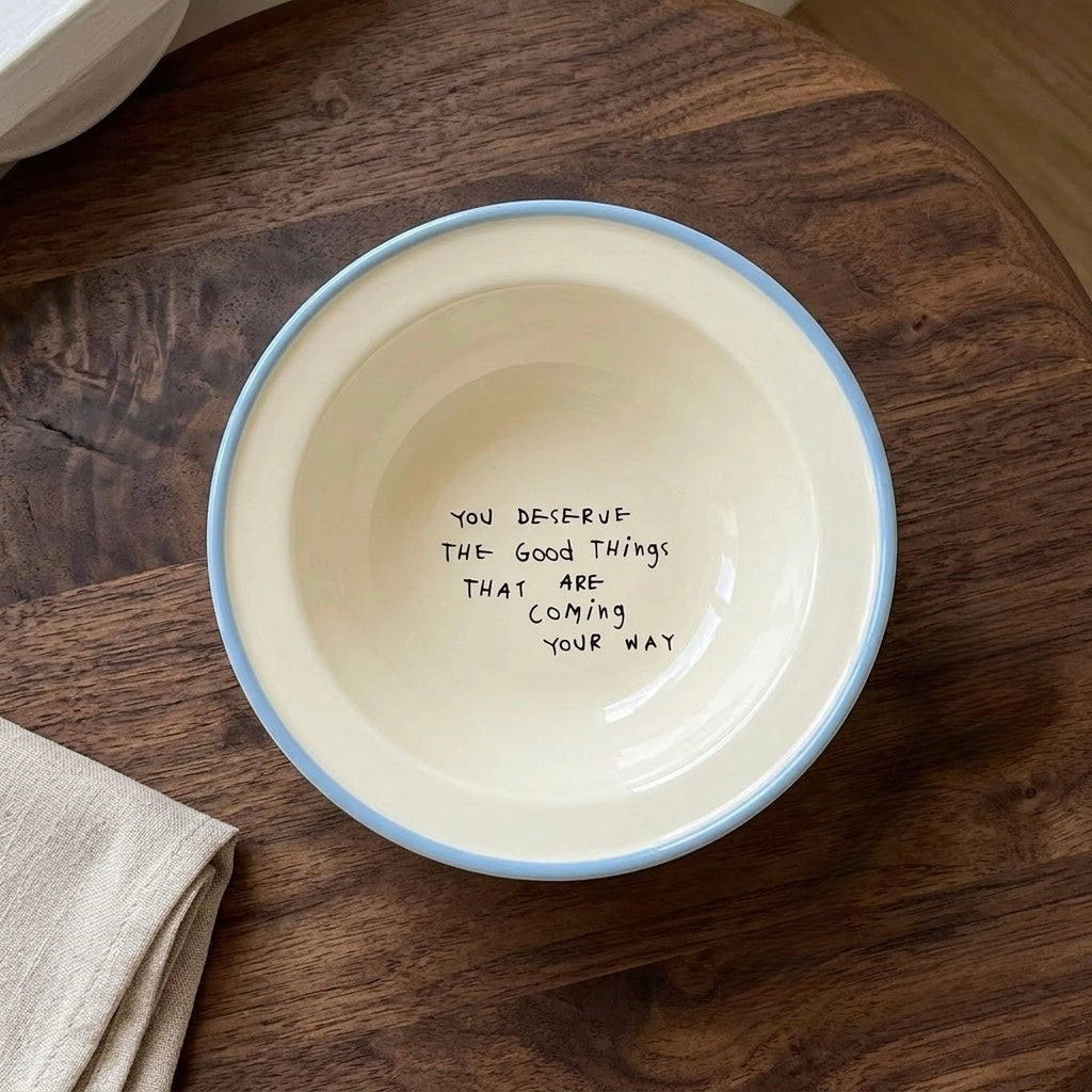 An off-white ceramic bowl with a light blue rim, featuring the handwritten text 'YOU DESERVE THE GOOD THINGS THAT ARE COMING YOUR WAY' inside, displayed on a dark wood tabletop.