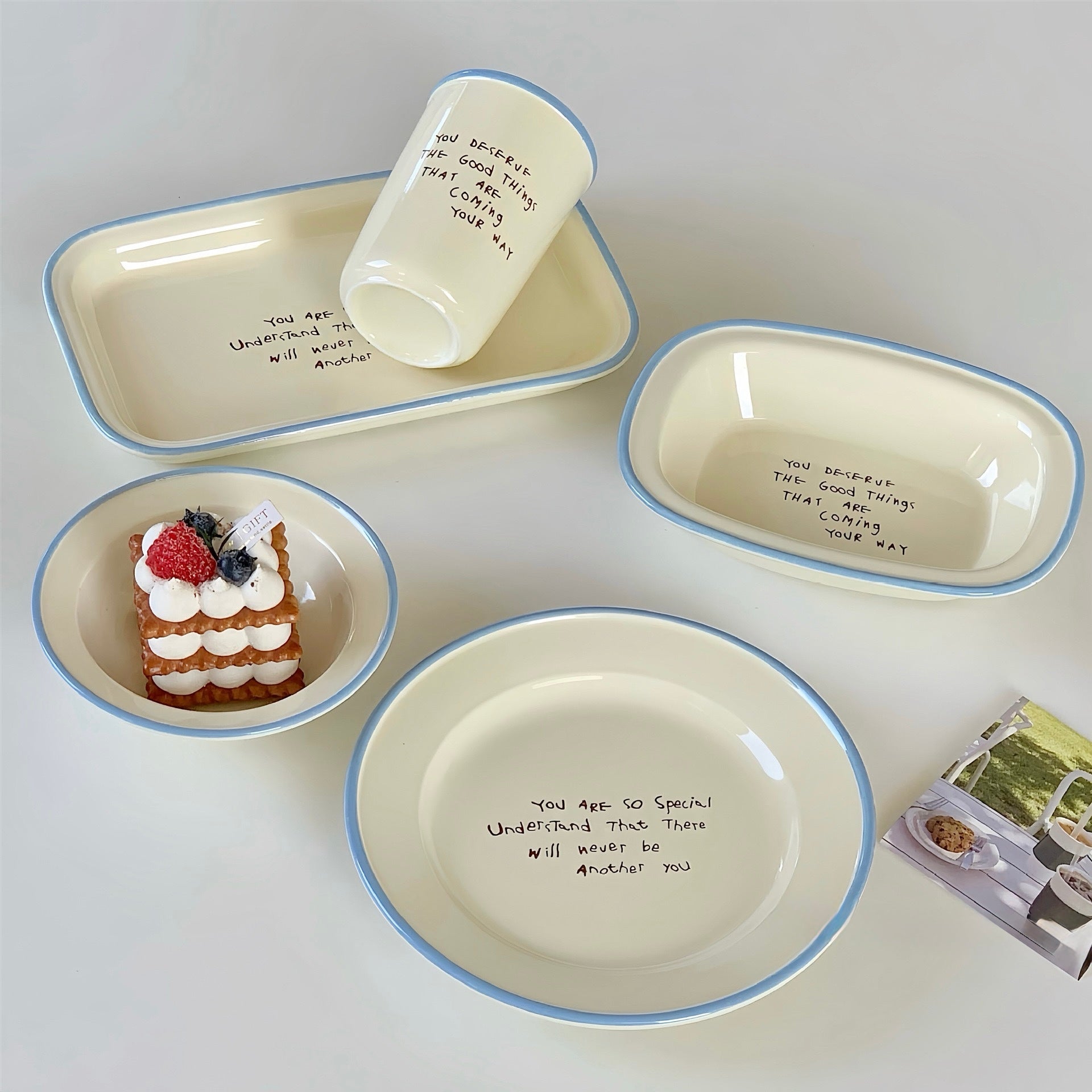 A set of cream-colored ceramic tableware with light blue rims, including a large plate, a rectangular tray, a small bowl with a dessert prop, and a cup, arranged on a light gray background.