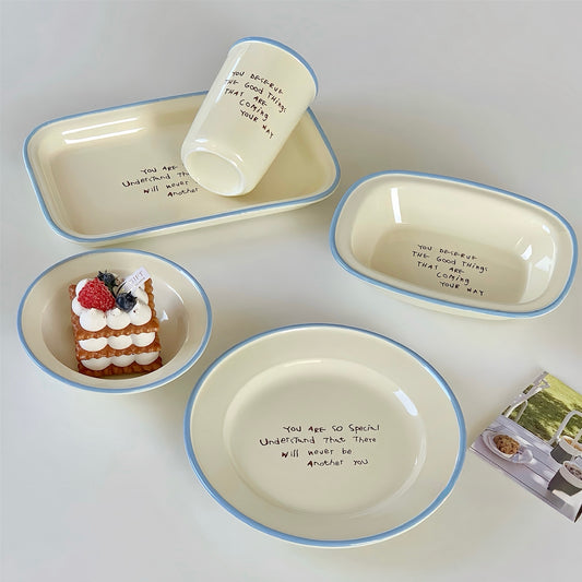 A set of cream-colored ceramic tableware with light blue rims, including a large plate, a rectangular tray, a small bowl with a dessert prop, and a cup, arranged on a light gray background.