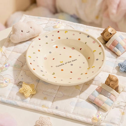 A round cream-colored ceramic bowl with a pattern of small stars, moons, and flowers, featuring the text 'Twinkle of the stars' printed inside. The bowl is styled on a quilted blanket with cute plush toys and decorative accessories.