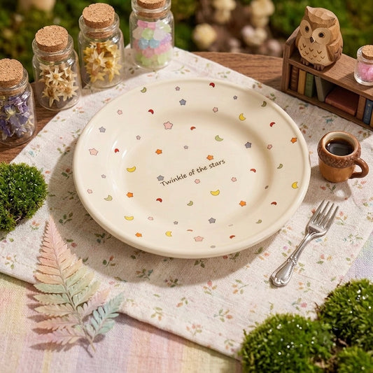 A round ceramic plate with a cream base decorated with tiny pastel star, moon, and heart motifs and the printed phrase 'Twinkle of the stars,' styled on a floral placemat with a fork, small cup, jars, moss, and an owl decoration in an outdoor garden setting.