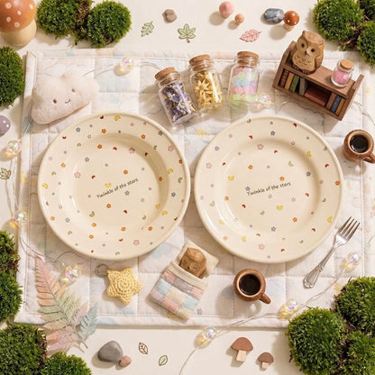 Two round cream-colored ceramic plates with small pastel floral and heart patterns and the text 'Twinkle of the stars' printed in the center, styled in a cozy flat-lay scene with miniature moss, cups, jars, and a small owl bookshelf.