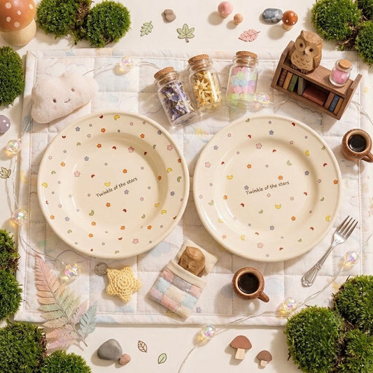 Two round cream-colored ceramic plates with small pastel floral and heart patterns and the text 'Twinkle of the stars' printed in the center, styled in a cozy flat-lay scene with miniature moss, cups, jars, and a small owl bookshelf.