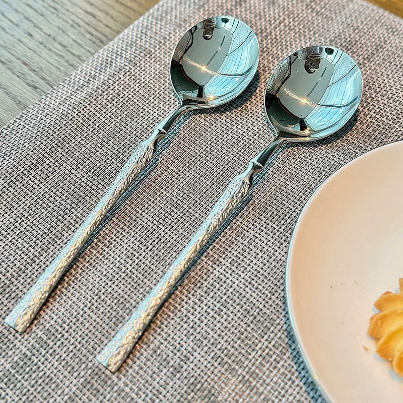 Two silver spoons on a textured placemat with a plate of food in the corner.