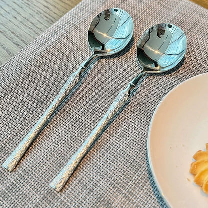 Two silver spoons on a textured placemat with a plate of food in the corner.