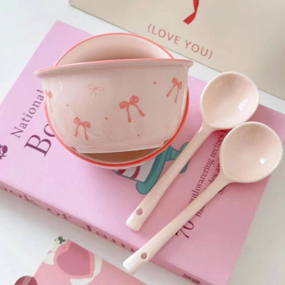 A set of ceramic bowls with a pink bow pattern, accompanied by matching spoons, displayed on a table with books in the background.