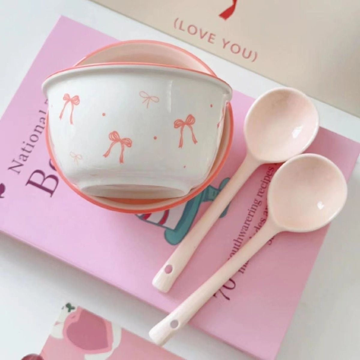 A set of ceramic bowls with a pink bow pattern, accompanied by matching spoons, displayed on a table with books in the background.