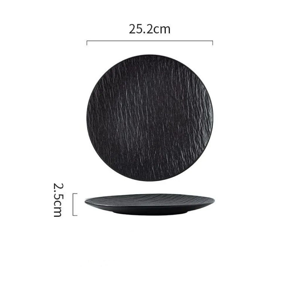 A black textured dinner plate with a matte finish, displayed on a gray surface with a napkin, wooden pepper grinder, and cutlery set to the side. A piece of food is served on the plate, showcasing its use in a dining setting.