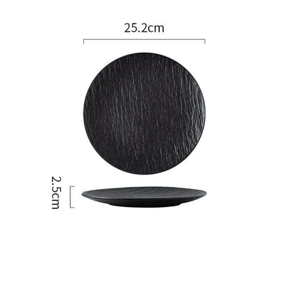 A black textured dinner plate with a matte finish, displayed on a gray surface with a napkin, wooden pepper grinder, and cutlery set to the side. A piece of food is served on the plate, showcasing its use in a dining setting.