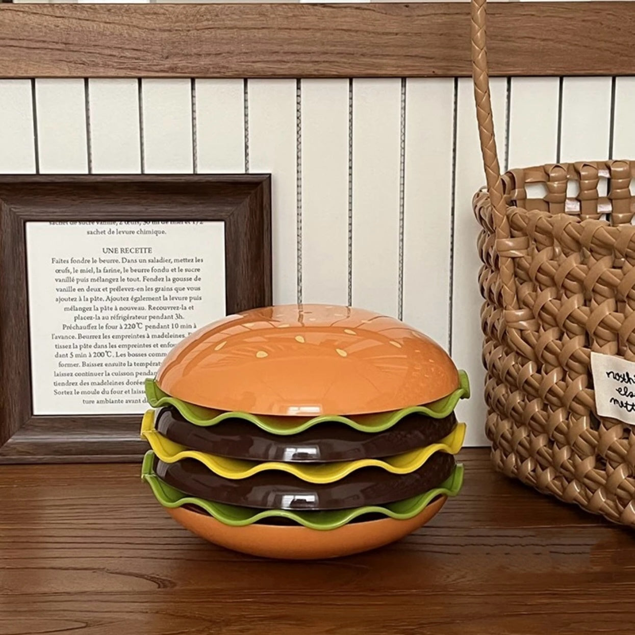 Hamburger-shaped bowl on a wooden surface with a framed recipe and woven basket in the background.