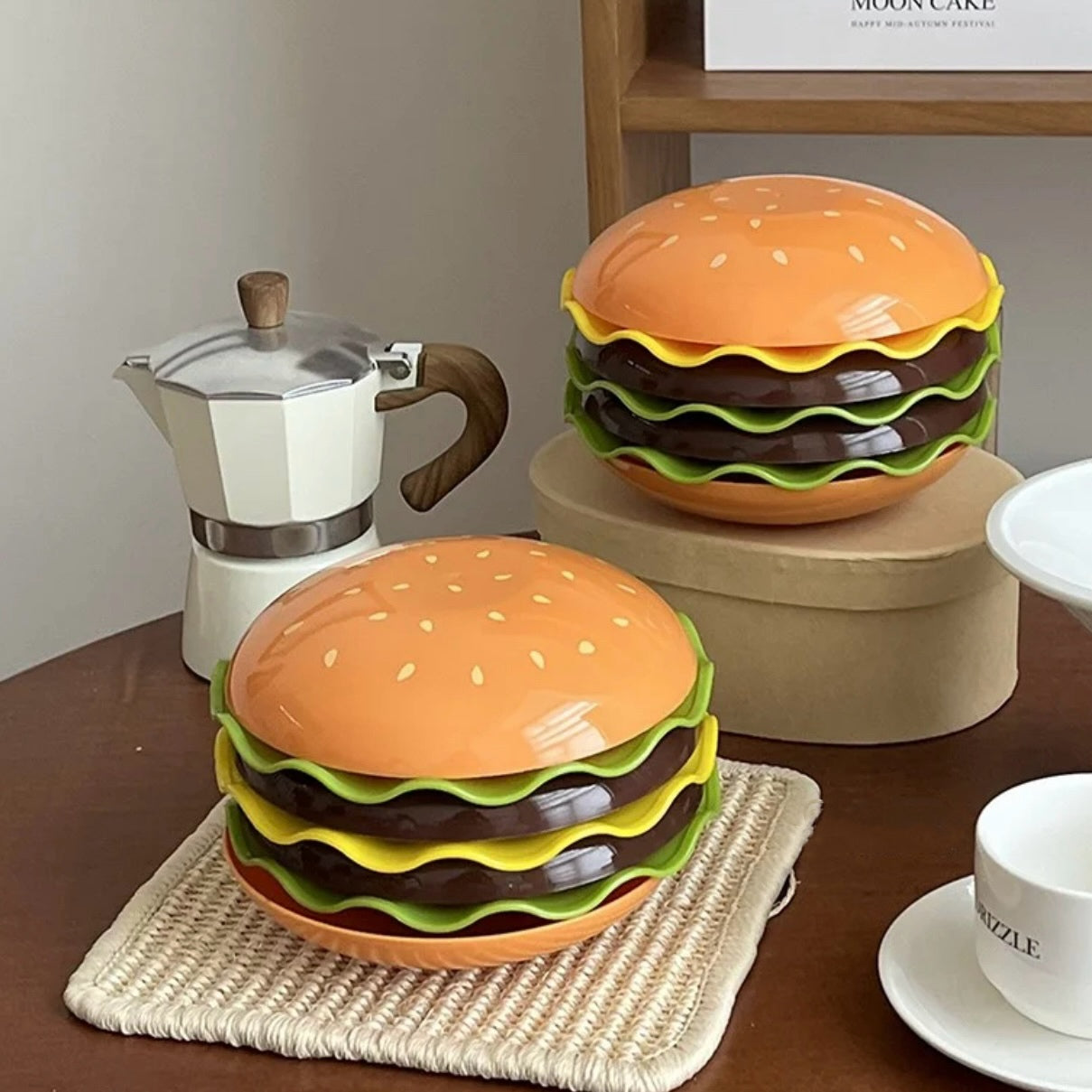 Two hamburger-shaped ceramic stacks on a table with a coffee pot and other items.