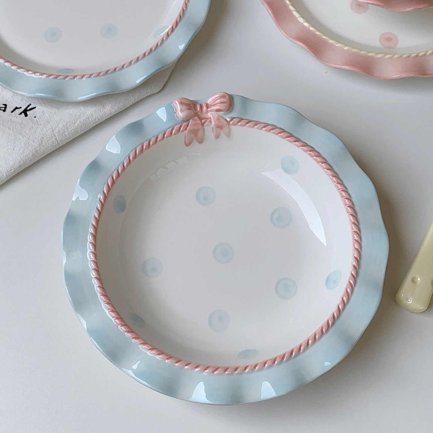 A set of ceramic tableware with a polka dot pattern and bow-knot detail, displayed in blue and pink colors.