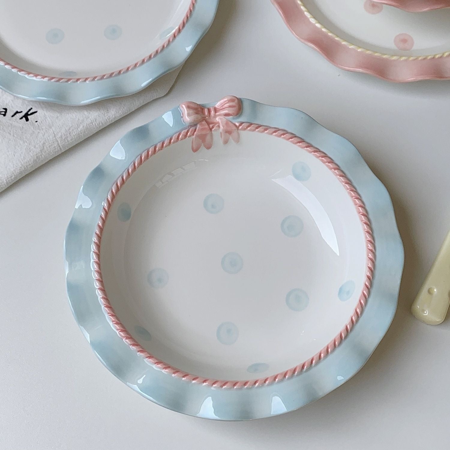 A set of ceramic tableware with a polka dot pattern and bow-knot detail, displayed in blue and pink colors.