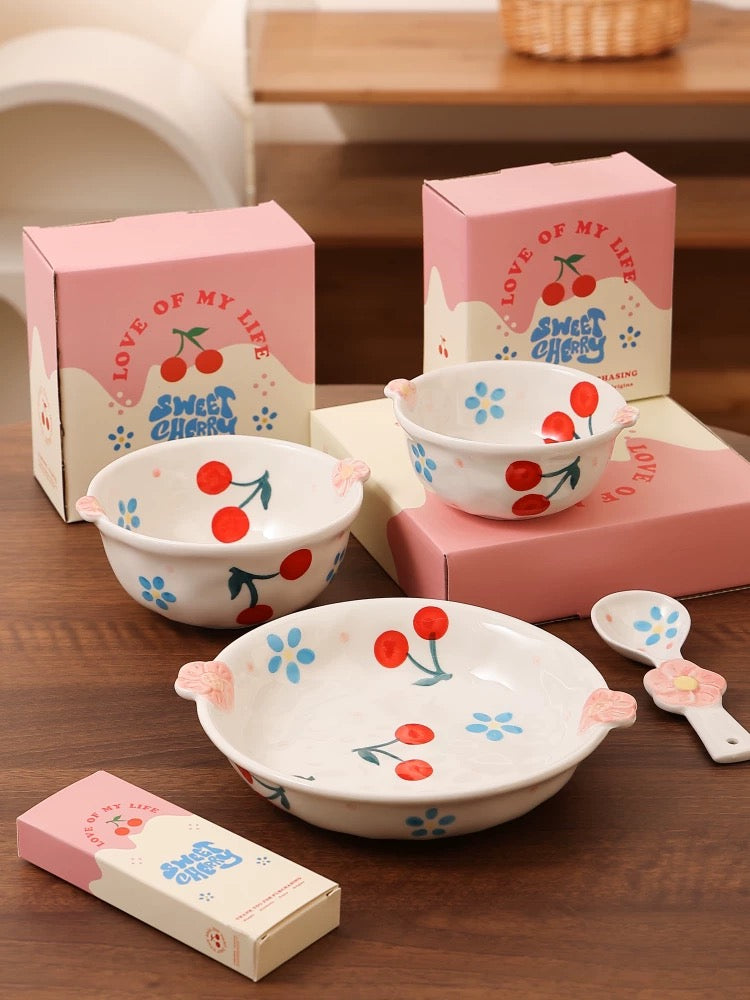 Set of ceramic bowls with cherry design on a wooden surface, surrounded by matching pink boxes.