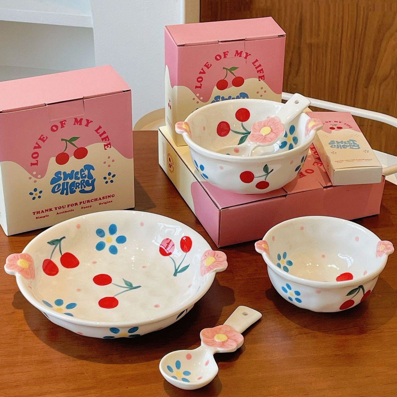 Ceramic bowls with cherry and flower design on a wooden surface, surrounded by packaging boxes.