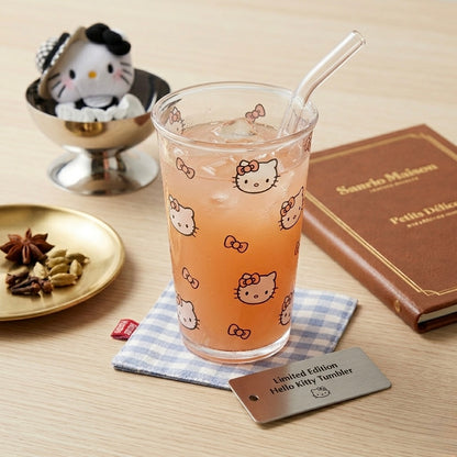 A clear glass tumbler with Hello Kitty face and bow prints, filled with an orange drink and a clear straw, styled on a wooden tabletop with a small Hello Kitty figurine, a brown book, and a metal tag reading 'Limited Edition Hello Kitty tumbler'.