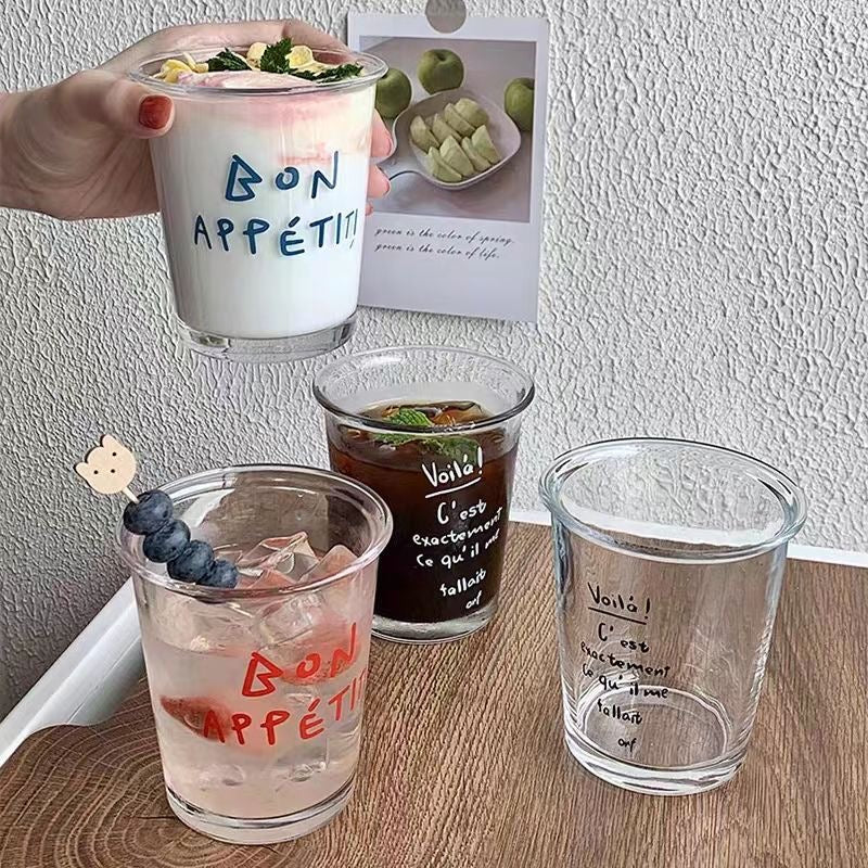 A set of transparent glass cups with handwritten-style French typography reading "Bon App¨¦tit" and "Voil¨¤." The cups are filled with different beverages, creating an aesthetic and cozy caf¨¦-style setting.