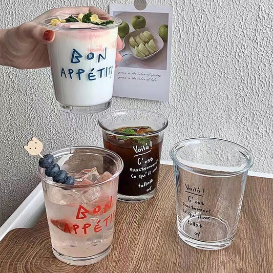 A set of transparent glass cups with handwritten-style French typography reading "Bon App¨¦tit" and "Voil¨¤." The cups are filled with different beverages, creating an aesthetic and cozy caf¨¦-style setting.