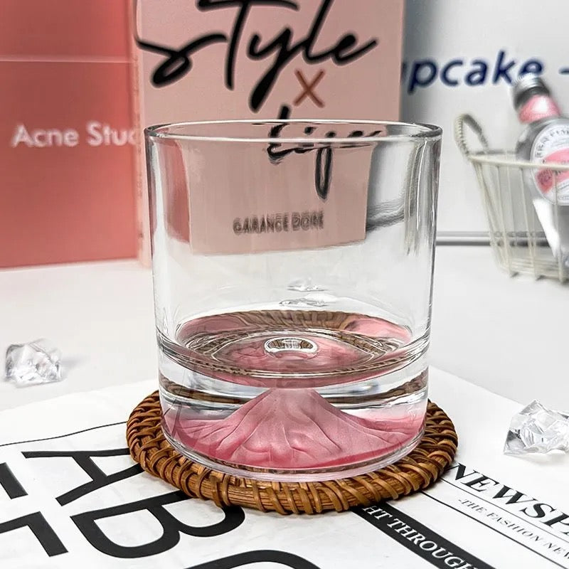 A stylish whiskey glass featuring a pink mountain design at the base, placed on a woven coaster with a modern aesthetic background.