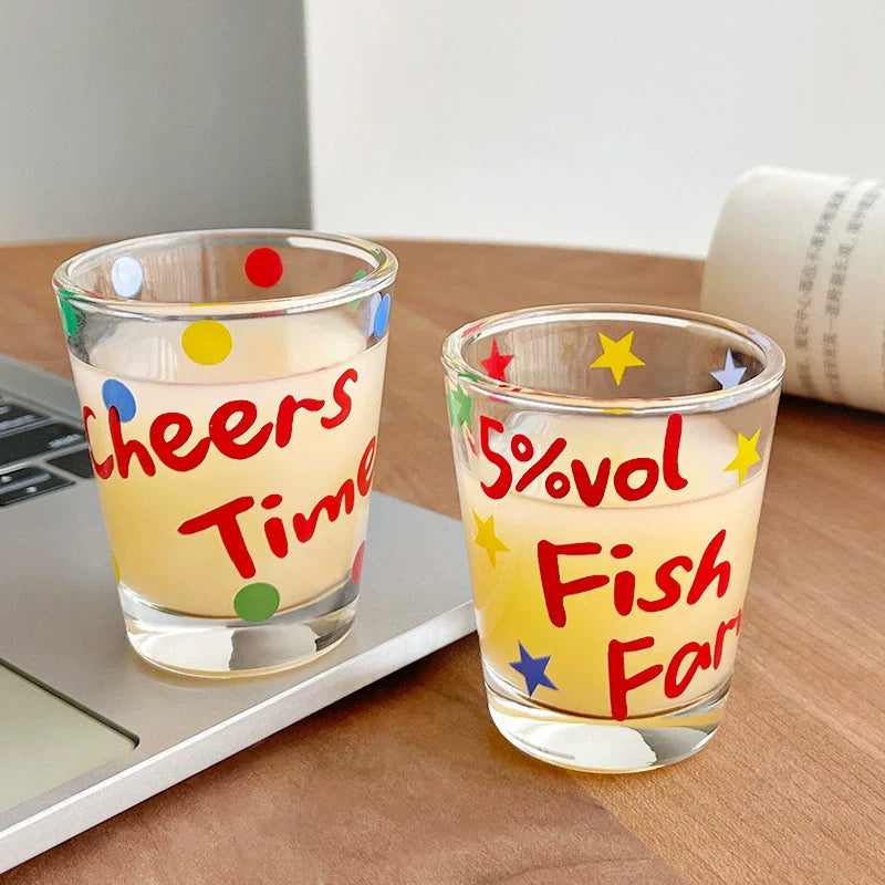 Two quirky shot glasses with playful text and colorful star and dot patterns.