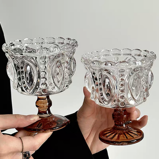 Two ornate glass candle holders with brown feet held by a person against a neutral background