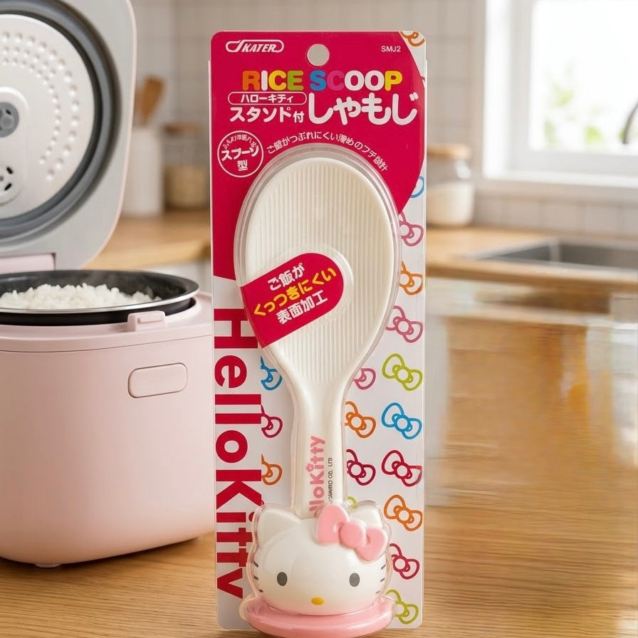 A packaged Tupper Hello Kitty rice scoop standing upright in front of a pink rice cooker on a wooden kitchen counter. The product is shown in its retail card packaging with Japanese text and Hello Kitty branding.