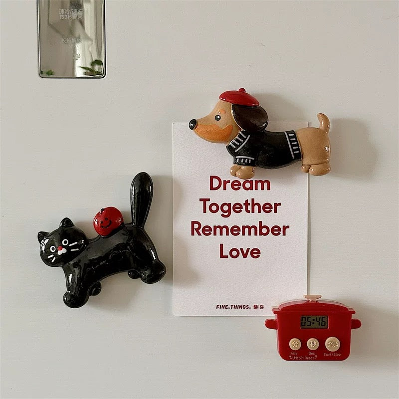 A black cat and a dachshund fridge magnet placed on a white surface, with the dachshund wearing a red beret and the black cat with a red apple on its back, next to a clock and above a postcard with a message.
