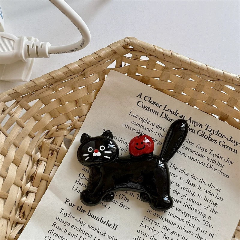 A black cat and a dachshund fridge magnet placed on a white surface, with the dachshund wearing a red beret and the black cat with a red apple on its back, next to a clock and above a postcard with a message.