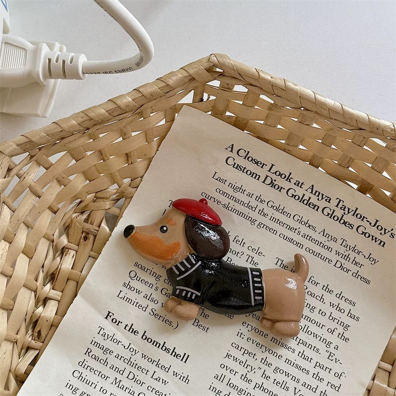 A black cat and a dachshund fridge magnet placed on a white surface, with the dachshund wearing a red beret and the black cat with a red apple on its back, next to a clock and above a postcard with a message.