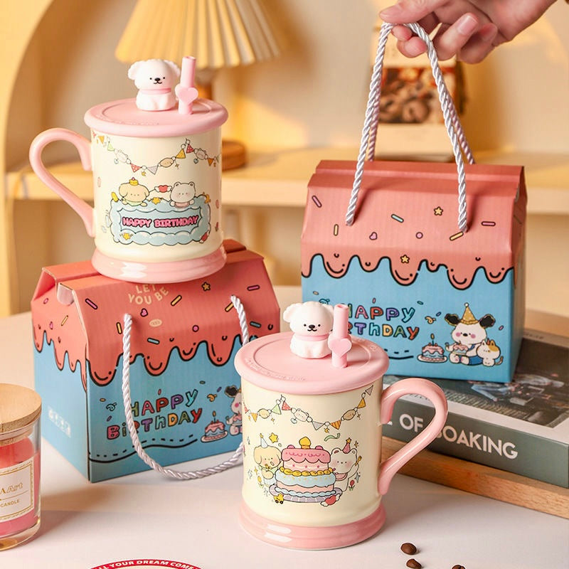 Two pink mugs with cartoon designs and lids, one held by a hand, on a surface with 'Happy Birthday' boxes.