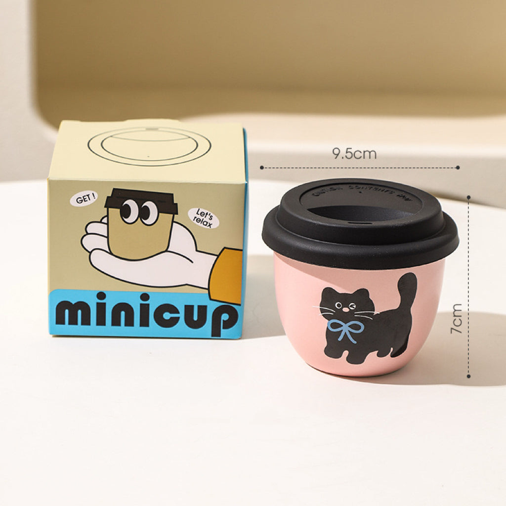 Stacked pink ceramic coffee cups with black cat designs, with a lid placed on one of the cups, displayed on a kitchen towel with a yellow and black box in the background.