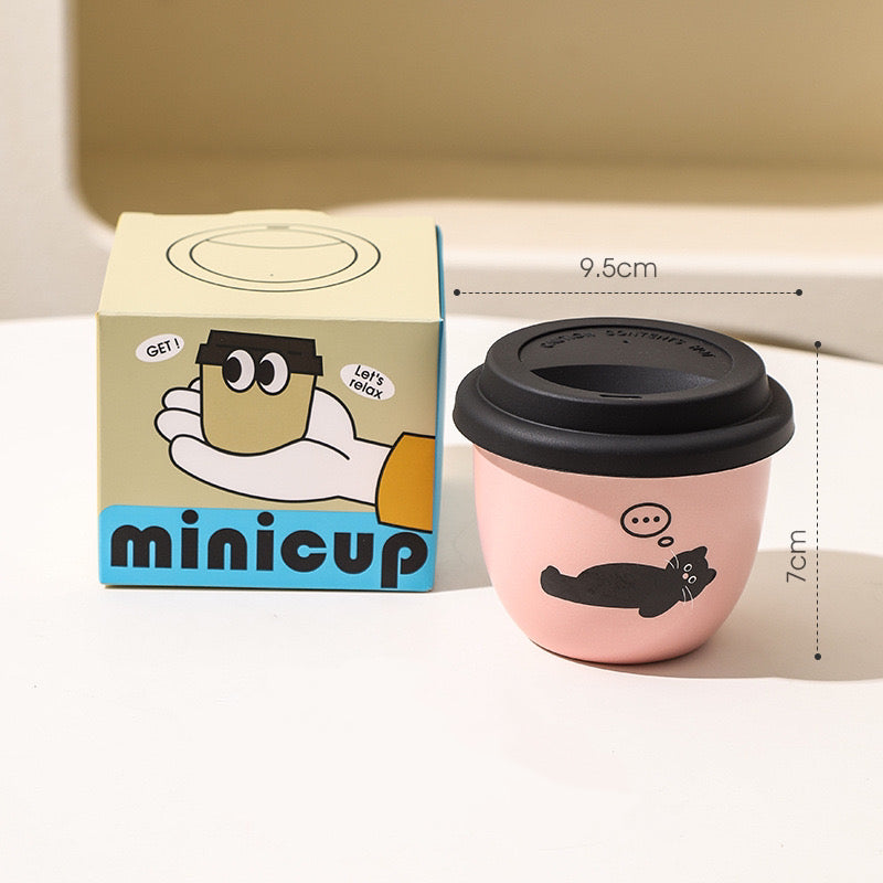 Stacked pink ceramic coffee cups with black cat designs, with a lid placed on one of the cups, displayed on a kitchen towel with a yellow and black box in the background.