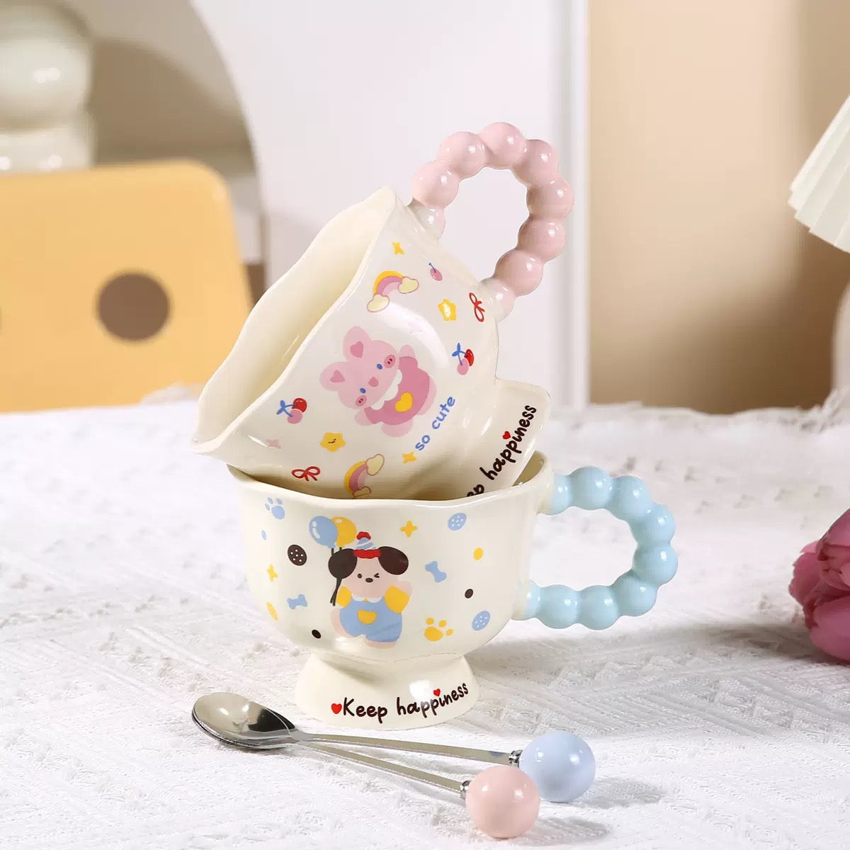 Two ceramic cups with cartoon designs, a spoon, and a teether on a soft surface.