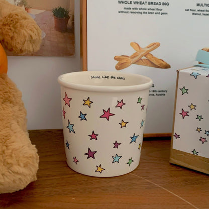 A handmade ceramic cup with a cute star pattern, displayed next to its packaging adorned with a blue ribbon.