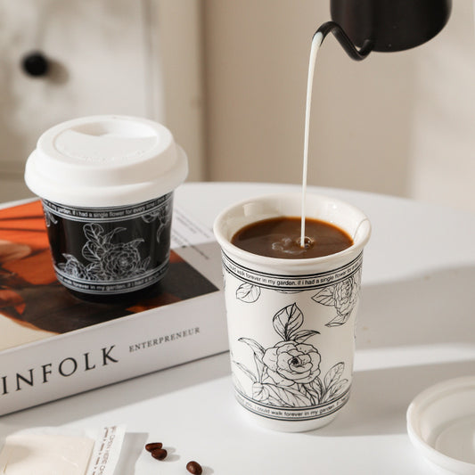 Coffee being poured into a white cup with black floral designs on a white surface.