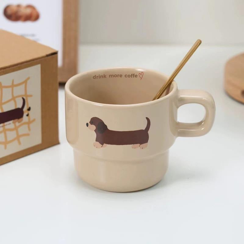 Two beige ceramic mugs with brown dachshund prints, stacked one inside the other, with a gift box visible in the background.