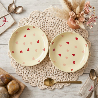 Two ceramic plates with heart patterns on a lace doily with utensils and flowers.
