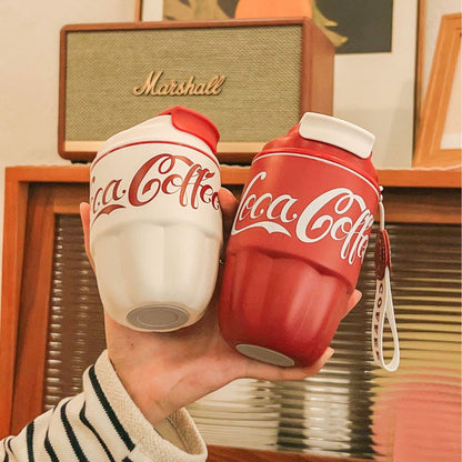 A stylish Coca Coffee Tumbler in red and white, inspired by the Coca-Cola bottle design, held in hand with a cozy background.